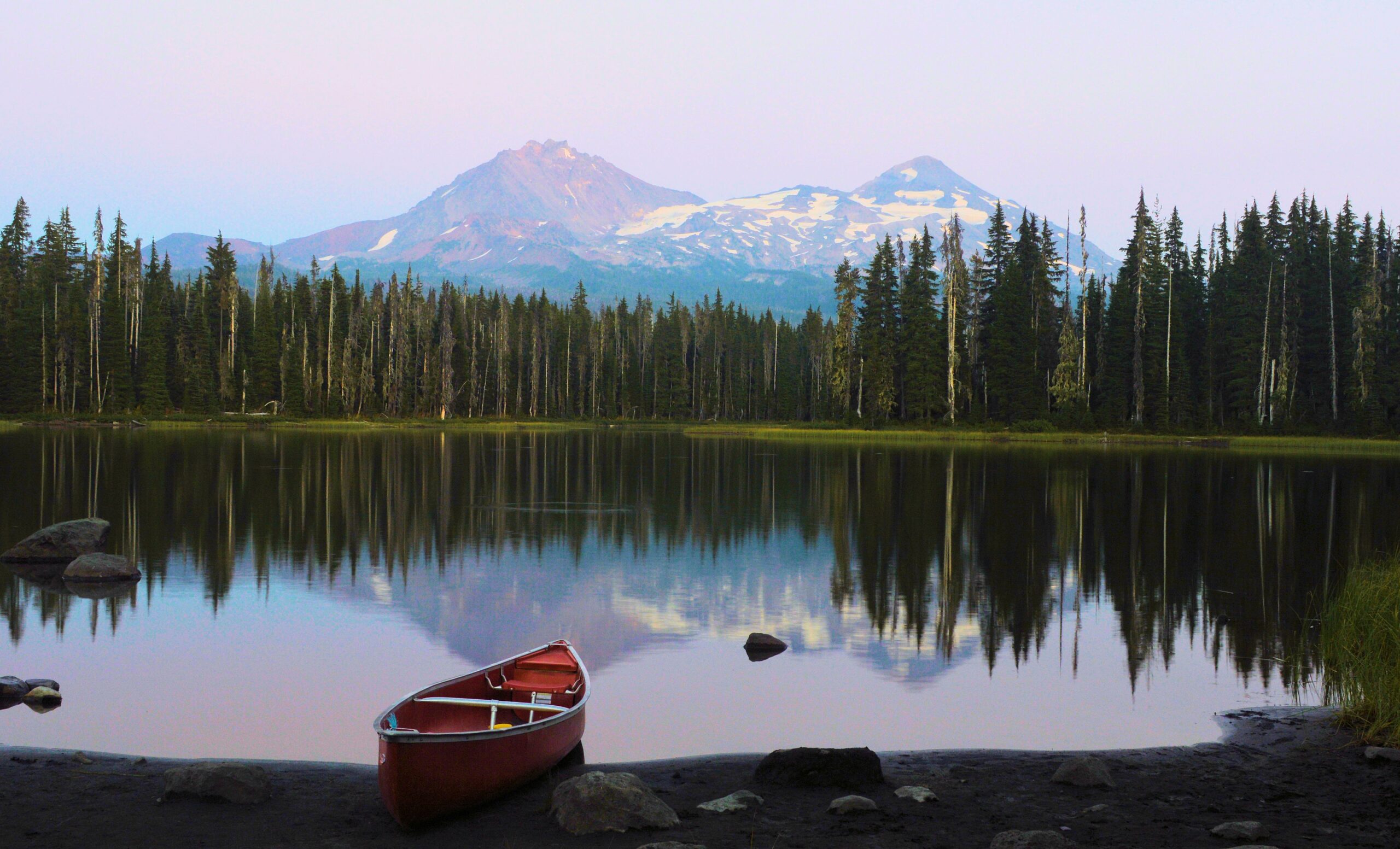 A canoe sits on the shore of a lake while a green forest rises up in the background, and beyond, snow covered mountains